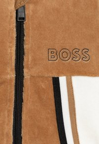 Giacca a zip in pile marrone con hardware nero, logo ricamato "BOSS" e accenti a strisce orizzontali in beige e bianco.