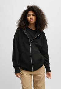 Felpa nera con zip realizzata in tessuto morbido, con tasca frontale, vestibilità comoda e cappuccio con cordino. Abbinata a pantaloni cargo beige.