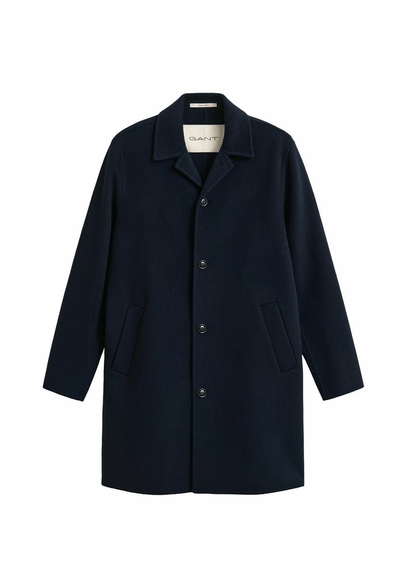 Long manteau bleu marine avec quatre boutons, deux poches latérales et un col rabattable, étiqueté GANT à l'intérieur du col.