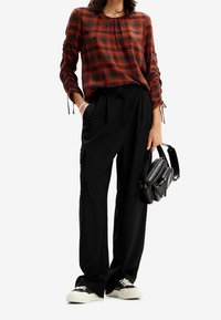 Donna che indossa una camicia a maniche lunghe a quadri rossi e neri, pantaloni neri a gamba larga, sneakers nere con suola bianca e tiene in mano una borsa nera.