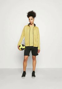 Κίτρινη φλις ζακέτα με μαύρες λεπτομέρειες, συνδυασμένη με μαύρες σορτς. Το άτομο κρατάει μια μαύρη και κίτρινη μπάλα ποδοσφαίρου. Απλός σχεδιασμός, casual εφαρμογή.