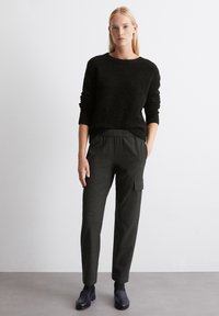 Maglione nero, con texture e scollatura rotonda, abbinato a pantaloni grigi scuri con tasca cargo e vita elasticizzata. Le scarpe nere completano il look.