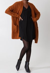 Cardigan orange surdimensionné en texture douce, avec deux poches avant. Porté sur une robe noire, associé à des bottines noires et des collants.