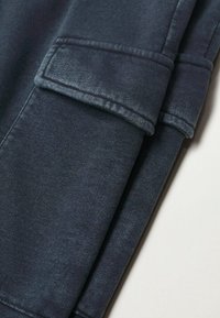 Tissu en denim bleu foncé avec une texture lisse. Présente un ourlet replié avec un détail de couture et une poche à rabat sur le côté.