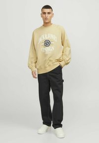 Sweatshirt beige à col rond avec logo et texte blancs, associé à un pantalon noir et des baskets blanches. Le tissu semble doux avec une coupe décontractée.