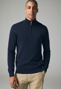 Pulover tricotat bleumarin cu guler înalt, prezentând un închidere cu fermoar pe un sfert și un mic accent cu logo. Textură fină și design ajustat.