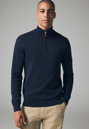 Navyblauer Strickpullover mit Stehkragen, ausgestattet mit einem Viertelreißverschluss und einem kleinen Logo-Detail. Glatte Textur und figurnahe Passform.