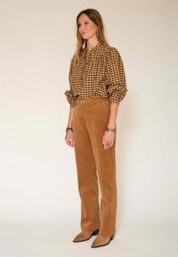 Blouse à carreaux marron avec des manches bouffantes, associée à un pantalon en velours côtelé marron. Le mannequin porte des bottes à bout pointu marron et des accessoires en argent.