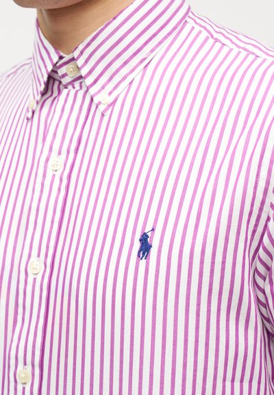 Κοντινό πλάνο ενός ατόμου που φοράει λευκό πουκάμισο με κάθετες μοβ ρίγες και μικρό κεντημένο λογότυπο ενός παίκτη πόλο πάνω σε άλογο.