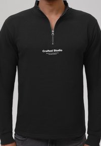 Svart zip-neck genser laget av et mykt materiale. Har en frontlogo i hvitt og tettsittende mansjetter, med et minimalistisk design.