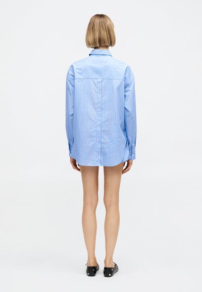 Άτομο με κοντά ξανθά μαλλιά στέκεται με την πλάτη προς το μέρος μας, φορώντας ένα μακρύ, ανοιχτό μπλε πουκάμισο με ρίγες και μαύρα παπούτσια με επίπεδη σόλα.