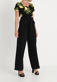 Pantalones anchos negros con una cintura ceñida por cinturón, combinados con una blusa negra de manga corta con un estampado floral de limones. Sandalias de tacón alto.