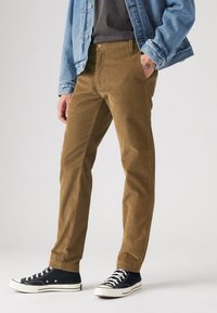 Pantalones de pana marrón con un corte recto, combinados con zapatillas de lona negras. El atuendo se completa con una chaqueta de mezclilla azul claro.
