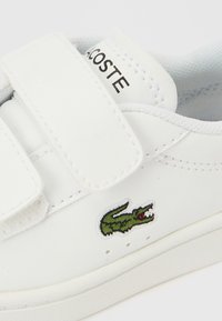 Λευκό αθλητικό παπούτσι με λουράκι βέλκρο, δερμάτινο υλικό, κεντημένο πράσινο λογότυπο κροκοδείλου και υφασμένη εσωτερική σόλα. Απλός σχεδιασμός, ελάχιστα μεταλλικά στοιχεία.