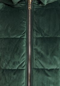 Grön pufferjacka med quiltat mönster, mjuk sammetstextur, svart dragkedja och huva. Guld detaljer längs dragkedjan.