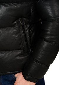 Schwarze Leder-Pufferjacke mit glatter Textur, Reißverschlusstasche auf der linken Seite und Manschettendetails, die die Ärmel betonen.