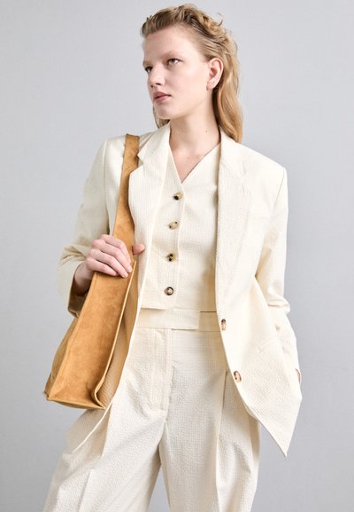Crèmekleurige blazer met gouden knopen, gecombineerd met een bijpassende top en hooggesneden broek. Draagt een tan kleurige suède tas.