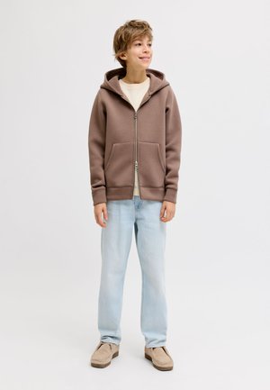 Ragazzo in piedi e sorridente, indossa una felpa marrone con cerniera, una camicia beige chiaro, jeans azzurri e scarpe beige su sfondo bianco.