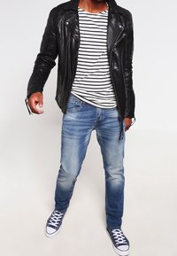 Chaqueta de cuero negra de motociclista, camisa a rayas blanca y marina, jeans de mezclilla deslavados y zapatillas marinas con suelas y cordones blancos.