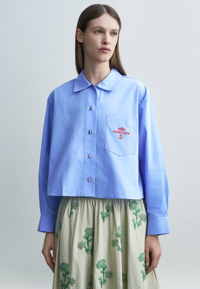 Femme portant une chemise courte boutonnée bleu clair avec le texte rouge "Yacht Club" sur la poche et une jupe beige à motifs floraux verts.