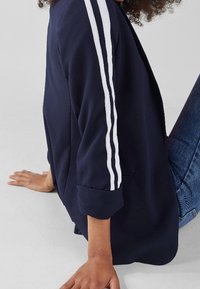 Marinblå blazer med vita ränder på ärmarna, med en avslappnad passform och rullade ärmavslut, tillverkad i ett mjukt tyg, kombinerad med blå jeans.