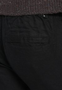 Juodos medžiagos kelnės matinio paviršiaus; turi kišenes su plokščiais, austais akcentais. Paviršius atrodo lygus, šiek tiek tekstūruotas.