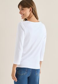 Hvid, langærmet bluse med rund halsudskæring, glat tekstur, parret med blå denim jeans med baglommer og en let falmning.