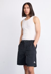 Man with long hair wearing a white sleeveless shirt and black cargo shorts with hands in pockets against a plain light background.