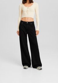 Top corto de canalé en crema con mangas largas y parte delantera con cordones, combinado con jeans de tiro alto de pierna ancha negros y zapatillas grises.