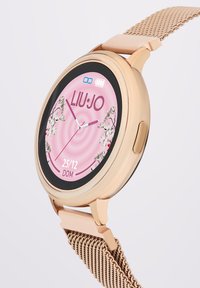 Guldtonet smartwatch med rund urskive, pink og blomsterdesign, sort bezel, datovisning og en mesh-metalrem.