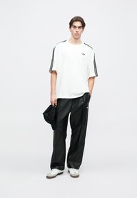 White short-sleeve shirt with black side stripes, paired with black loose-fitting pants. White shoes and a black cap held in hand.