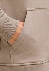Sudadera beige con bolsillo y una mano parcialmente visible. La textura es suave, con puños acanalados y un detalle de costura limpio a lo largo del bolsillo.