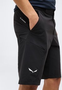 Άτομο που φοράει μαύρα σορτς DuraStretch με το χέρι στην πλαϊνή τσέπη, που διαθέτει τσέπη στο μηρό με φερμουάρ και λευκό λογότυπο κοντά στο τελείωμα.