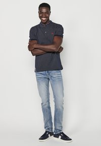 Jūrinė polo marškinėliai su tekstūruotu raštu, trumpomis rankovėmis ir apykakle; derinami su šviesiai mėlynomis džinsomis ir jūriniais sportbačiais su baltais akcentais.