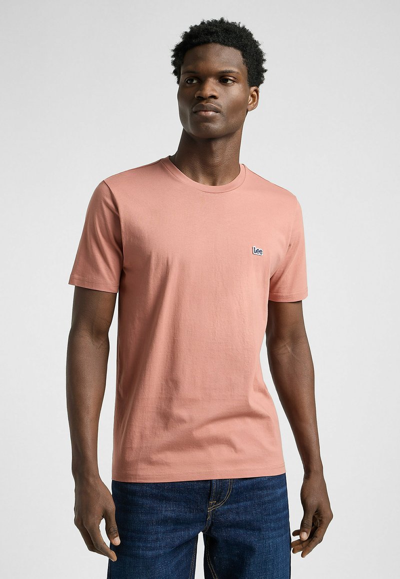 Bawełniana koszulka w delikatnym odcieniu koralowym, krótki rękaw, okrągły dekolt, z małym niebieskim logo "Lee" na piersi, zestawiona z ciemnoniebieskimi dżinsami.