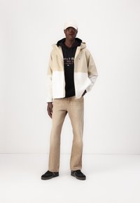 Hooded jacket in beige and white, black sweatshirt underneath, light denim pants, black sneakers, and a light cap. Simple design, casual style.