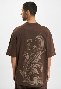 Brun bomulds t-shirt med et stort ornamentalt design og teksten "DAYS OF OLD" trykt i en lys farve på ryggen. Korte ærmer.