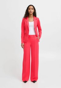 Helles pinkes Anzugset mit einem taillierten Blazer mit einem einzigen Knopf und weiten Hosen. Der Stoff wirkt glatt mit minimalen Details.
