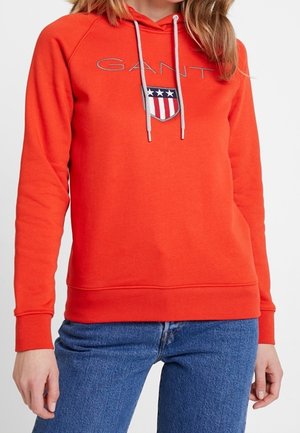 Sweat-shirt rouge avec capuche, arborant un logo et un design de blason sur la poitrine. Cordon de serrage gris, poignets et ourlet côtelés. Matière en mélange de coton.