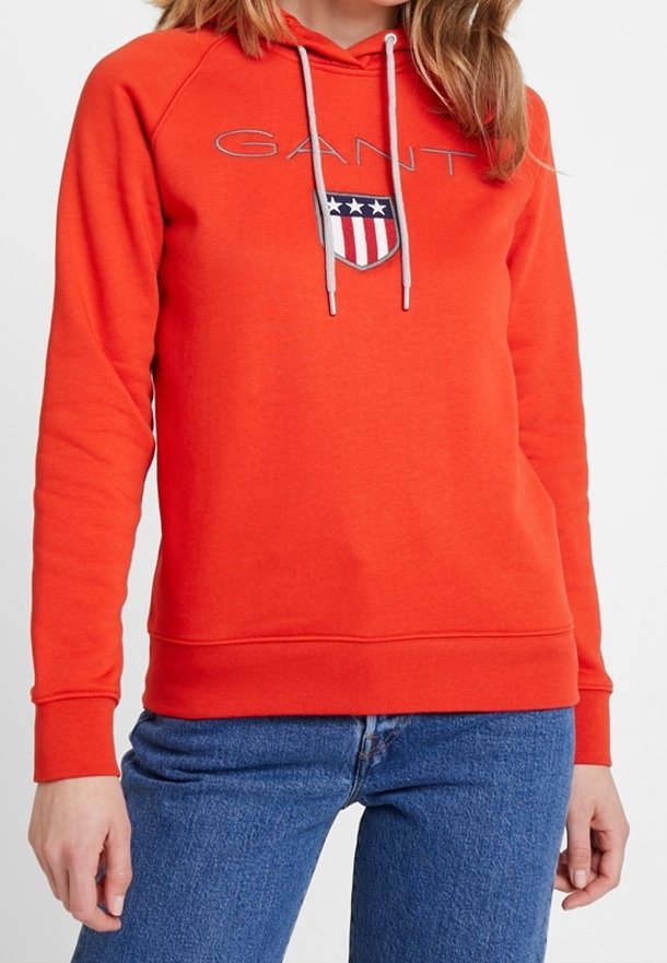 Sweat-shirt rouge avec capuche, arborant un logo et un design de blason sur la poitrine. Cordon de serrage gris, poignets et ourlet côtelés. Matière en mélange de coton.