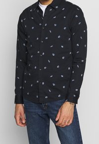 Camicia nera a maniche lunghe con colletto a bottone, caratterizzata da un motivo blu e bianco su tutta la superficie. Tessuto morbido, vestibilità regolare.