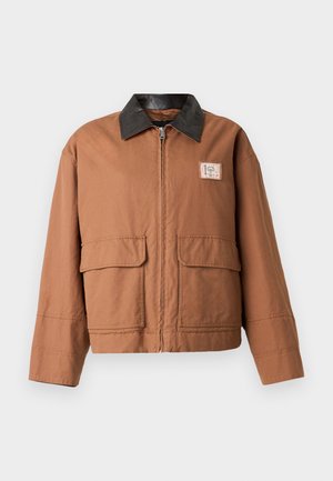 Bruine jas met rits en zwarte kraag, twee klepzakken aan de voorkant, lange mouwen en een klein rechthoekig embleem aan de rechterkant van de borst.