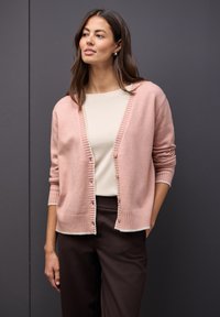 Růžový vlněný cardigan s žebrovanými detaily, zapínáním na knoflíky a dlouhými rukávy, zkombinovaný se smetanovým trikem a tmavě hnědými kalhotami.