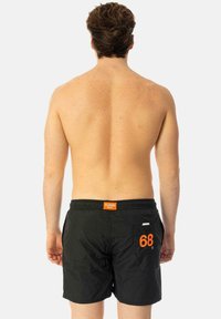 Costume da bagno neri con vita elasticizzata, dotati di un'etichetta arancione e una tasca laterale con il numero "68" in arancione. Materiale leggero.