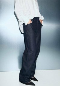 Jeans de mezclilla oscura de pierna ancha con una textura lisa y un brillo sutil, combinados con una camisa blanca holgada y zapatos negros de punta.