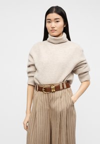 Γυναίκα με μακριά μαύρα μαλλιά που φοράει μπεζ πουλόβερ με ζιβάγκο, ψηλόμεσο ριγέ παντελόνι και καφέ ζώνη με χρυσή αγκράφα.