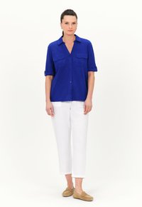 Camicetta blu a maniche corte con due tasche sul petto, abbinata a pantaloni bianchi a vita alta e scarpe slip-on color cammello.