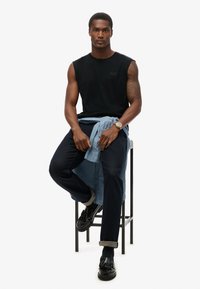 Camiseta sin mangas negra, chaqueta de mezclilla atada a la cintura, jeans oscuros, mocasines negros con borlas, sentado en un taburete de metal, fondo neutro.