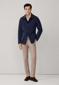 Marineblau gesteppte Jacke mit braunem Kragen, zwei Fronttaschen, Knopfverschluss, kombiniert mit beigen Hosen und braunen Schuhen. Glatte Textur.