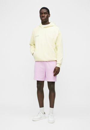 Junger Mann steht, trägt einen hellgelben Kapuzenpullover, hellrosa Shorts, weiße Socken und weiße Sneakers vor einem schlichten weißen Hintergrund.